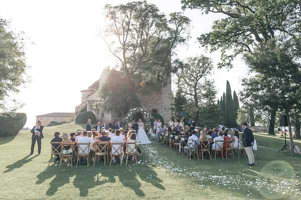 An outdoor wedding ceremony taking place on a lawn in front of ancient stone ruins, likely a medieval tower or château remnant in the French countryside. Approximately 60-80 guests are seated in wooden cross-back chairs arranged in rows on either side of a petal-lined aisle, with white flower petals scattered along the ground leading to the altar. The couple stands at the front beneath a large circular floral arch decorated with white and green foliage, with an officiant to one side. The bride is wearing a white gown and the scene is shot as a wide aerial-perspective shot taken from an elevated angle, capturing the full layout of the ceremony space and long shadows cast by late afternoon sunlight. Potential venue feature image.