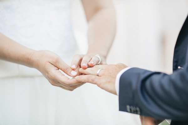 Close-up detail shot of a ring exchange moment during a wedding ceremony, showing the bride placing a plain gold band onto the groom's finger. The bride is wearing a white dress and has coral-painted fingernails, while the groom is dressed in a navy blue suit jacket. The background is softly blurred, revealing light neutral tones consistent with an indoor or covered outdoor ceremony setting.