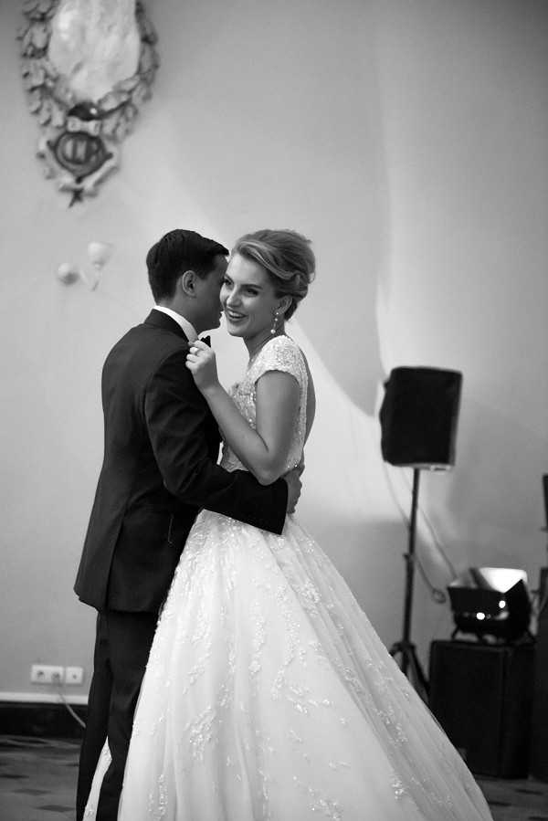A black-and-white portrait-style shot of a couple sharing their first dance indoors during a wedding reception. The groom wears a dark suit with a bow tie, and the bride wears a full ballgown with a lace-embellished bodice, cap sleeves, and a voluminous skirt; her hair is styled in an updo and she wears drop earrings. The bride is smiling and facing the camera while the groom leans close to her. The setting appears to be a formal interior room with an ornate decorative wall medallion or coat of arms mounted above them, and DJ or audio equipment is visible in the background to the right. The image has strong contrast with soft highlights on the bride's dress and face.