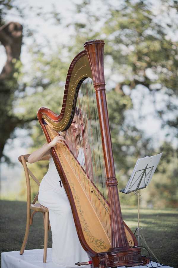 A female harpist performs outdoors at a wedding, seated on a wooden cross-back chair behind a large concert harp with a warm mahogany finish and gold-painted soundboard detailing. She is wearing a white dress and smiling while playing. A black music stand with sheet music is positioned to her right. The setting appears to be a garden or grounds of an outdoor venue, likely during a ceremony or cocktail hour. The portrait-style shot is taken at a slight angle, with the harp partially obscuring the musician, and the background is softly out of focus.
