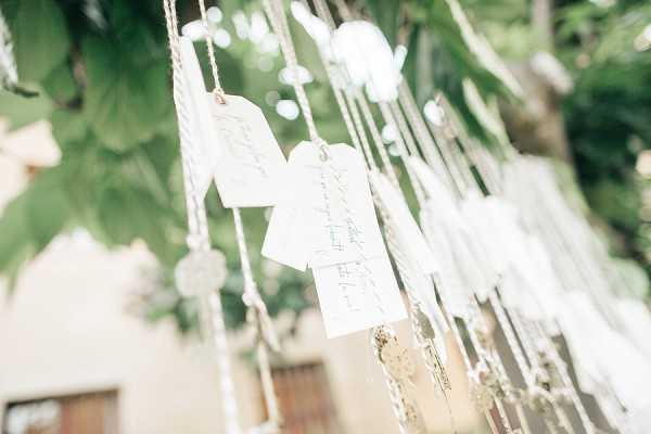 Close-up detail shot of a wedding seating chart or escort card display, where individual white paper tags with handwritten guest names are hung from strings or twine suspended from a tree outdoors. The tags are interspersed with small decorative crystal or acrylic disc accents threaded along the cords. The background is softly out of focus, showing green foliage and a glimpse of a building facade, suggesting a courtyard or garden setting. The overall styling is light and airy with a rustic-meets-delicate aesthetic, using natural twine and handwritten cards.