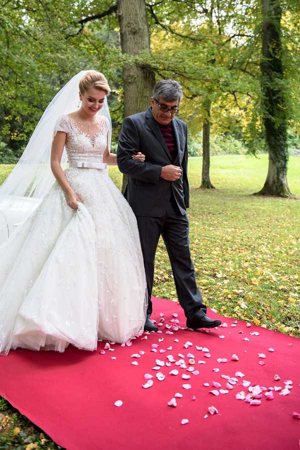 A bride is walking down a red carpet aisle during an outdoor ceremony, escorted by an older man — likely her father — who is holding her arm. The bride wears a full ballgown with a lace and embellished bodice featuring cap sleeves, a satin bow waistband, and a full tulle skirt, paired with a long cathedral-length veil; her blonde hair is styled in an updo. The red carpet aisle is scattered with pink rose petals as decoration. The setting is a garden or parkland with mature trees, and the shot is a medium portrait taken at ground level capturing both figures from approximately the knees up.
