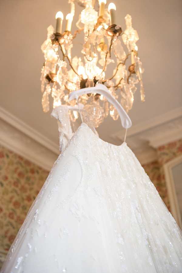 A getting-ready detail shot of a white ball gown-style wedding dress hanging from a crystal and gilt chandelier inside a chateau or manor house interior. The dress features all-over beading or lace appliqué with a full skirt and is hung on a white satin-ribbon hanger. The room is decorated with floral-patterned wallpaper in warm pink and green tones, and the chandelier's candle-style bulbs are lit, casting a warm amber glow. The image is taken from a low angle looking up toward the chandelier, emphasizing the volume of the gown's skirt in the foreground.