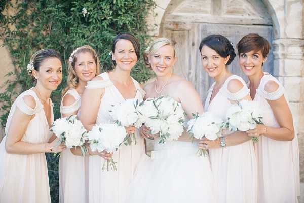 A bridal party portrait featuring a bride and five bridesmaids posing closely together outdoors in front of a stone wall with a wooden door. The bride wears a white strapless gown and a pearl necklace, while the five bridesmaids are dressed in matching blush/champagne floor-length gowns with cold-shoulder strap detailing. All six women hold round bouquets of white peonies with green foliage. The bride and several bridesmaids have small white floral hair accessories. The overall color palette is soft white and blush, giving the group a cohesive, classic look. This is a close-up group portrait shot from approximately waist height.
