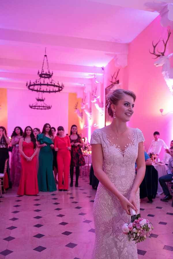 The bride is preparing to toss her bouquet during the indoor wedding reception, standing on a black-and-white diamond-patterned marble floor with a group of approximately ten female guests lined up behind her in anticipation. The venue is a formal ballroom or chateau reception room featuring wrought-iron candelabra-style chandeliers, mounted deer antler wall decor, and white walls bathed in deep pink and magenta uplighting. The bride wears a fitted, heavily beaded and lace cap-sleeve gown with a deep V-neckline, pearl drop earrings, and a matching necklace; she holds a small bouquet of blush and pink roses with white peonies. The guests behind her are dressed in a mix of vibrant formal wear including teal jumpsuits, red gowns, and coral and jewel-toned dresses. The shot is a medium portrait taken from a low angle slightly in front of the bride, with the assembled guests softly out of focus in the background.