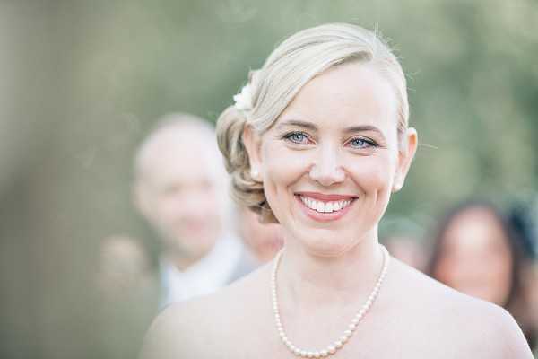 Close-up portrait of a bride smiling at the camera during what appears to be an outdoor ceremony or reception. She has blonde hair styled in a low updo with a small white floral hair accessory, and wears a pearl necklace and a strapless dress. Her makeup is natural with defined eyes. In the soft-focus background, a man in a light-colored shirt and at least one other guest are partially visible. The image has a light, airy feel with a shallow depth of field that keeps the bride sharply in focus against the blurred green background.