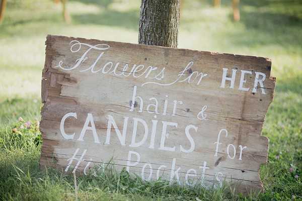 Close-up shot of a rustic wooden sign made from reclaimed pallet boards, propped against a tree trunk in an outdoor garden setting. The sign is painted in white hand-lettered text reading 'Flowers for HER hair & CANDIES for His Pockets,' directing guests to wedding favors or activity stations. The weathered, distressed wood and hand-painted lettering reflect a rustic, country-style wedding aesthetic. A second wooden sign is partially visible in the soft-focus background.