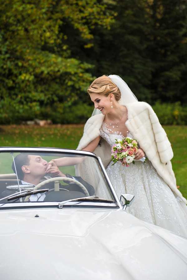A couple portrait taken outdoors, with the groom seated in a vintage white convertible car kissing the bride's hand through the open window. The bride leans over the car door, smiling, wearing a heavily embellished ball gown with a cathedral-length veil, a cream fur stole draped over her shoulders, and carrying a bouquet of blush pink roses, white ranunculus, and small mixed blooms. The groom is dressed in a dark suit. The styling has a classic, old-Hollywood aesthetic. Medium portrait shot with a shallow depth of field, taken in a garden or estate setting.
