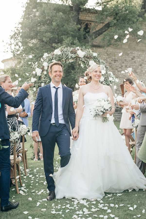 A newly married couple walks back up the aisle during their outdoor ceremony recessional, with approximately 20 guests on either side tossing white flower petals into the air. The groom wears a navy blue suit with a navy tie and light blue shirt, while the bride wears a strapless white ball gown with a layered tulle skirt and carries a bouquet of white roses and greenery. A circular floral arch made of greenery and white roses frames the couple from behind, positioned in front of a stone building partially obscured by trees. Guests are dressed in summer attire in neutral and pastel tones, and wooden folding chairs line the aisle. The image is a medium full-length portrait shot taken in bright, soft natural daylight.