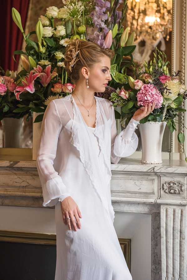 A bridal portrait shot indoors, with a bride posing beside an ornate white marble fireplace mantel. She wears a white sheer long-sleeve gown with lace trim detailing and a ruffled front, accessorized with drop pearl earrings, a delicate pendant necklace, and a gold hair accessory in her upswept hair. She lightly touches a large pink hydrangea cluster held in a white ceramic vase, which is part of an abundant floral arrangement on the mantel featuring pink oriental lilies, lavender delphiniums, white lisianthus, and garden roses in mixed pink, white, and purple tones. A crystal chandelier and a gold-framed mirror are visible in the background, suggesting a classic French interior setting. The composition is a three-quarter portrait with soft, warm indoor lighting and a slightly blurred background that draws focus to the bride.