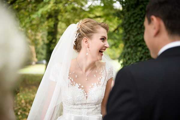 An outdoor portrait capturing a candid moment between a bride and groom, with the bride laughing openly toward the groom whose back is partially visible in the foreground. The bride wears a white lace gown with a sheer illusion neckline decorated with floral appliqués and cap sleeves, paired with a long veil and a decorative hair accessory; she also wears pearl drop earrings. The setting appears to be a garden or estate grounds with ivy-covered walls visible in the background. The shot is a close-up portrait with a shallow depth of field, keeping focus on the bride's expression.