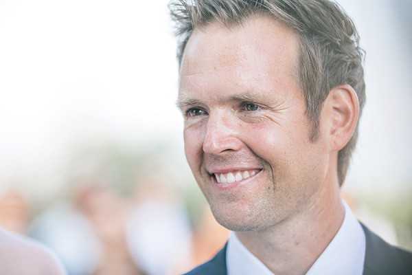 Close-up portrait of a smiling man, likely the groom, wearing a dark suit jacket and white dress shirt. The shot is tightly framed on his face, with a heavily blurred outdoor background suggesting a bright, open-air setting with other guests visible in soft focus. The image is sharp on his features with a shallow depth of field, giving it a candid, documentary feel.