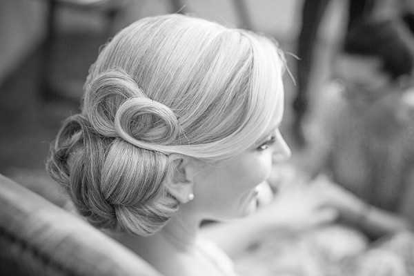 Black-and-white close-up portrait of a bride during the getting-ready phase, focusing on her styled updo hairstyle. The hair is swept into a low chignon with a rolled, sculpted detail at the nape of the neck and soft, swept-back sections framing the face. She is wearing a small pearl or stud earring and appears to be smiling slightly, looking to one side. The image has soft contrast with bright highlights on the hair, giving it a polished, classic feel.