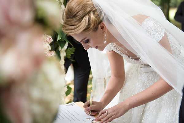 The bride is signing the marriage register during an outdoor ceremony, leaning forward over a document and holding a gold pen. She wears an ivory lace gown with cap sleeves and beaded detailing, paired with a long tulle veil and drop pearl earrings, with her blonde hair styled in an updo. A figure in a dark suit is partially visible behind her, and blush pink and cream floral arrangements are softly out of focus in the foreground. The shot is a close-up portrait with shallow depth of field, emphasizing the signing moment.