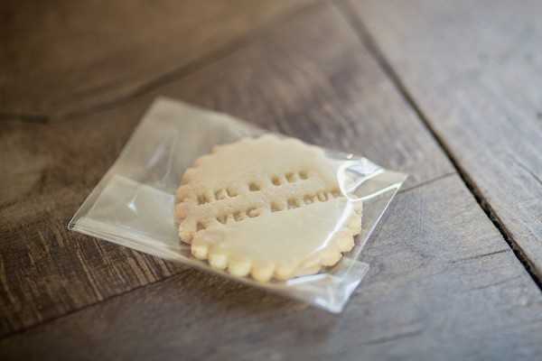 Close-up detail shot of a round, scallop-edged shortbread-style sugar cookie in a clear cellophane favor bag, placed on a rustic wooden surface. The cookie is ivory/cream in color and has text stamped or embossed into it, partially legible as what appears to read 'avec amour' or similar French wording, suggesting it is a personalized wedding guest favor. The composition is a shallow depth-of-field detail shot with soft, natural lighting highlighting the texture of the cookie and the wood grain beneath.