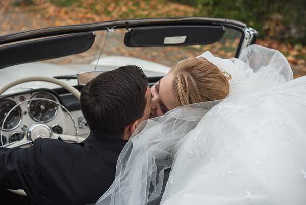 The bride and groom are sharing a kiss inside a vintage white Mercedes-Benz convertible, shot from behind and slightly above in a close portrait composition. The groom is wearing a dark navy or black suit jacket, and the bride's full white tulle veil and voluminous ball gown skirt cascade over the cream leather interior of the classic car. The vintage Mercedes dashboard and steering wheel are clearly visible, adding a retro styling detail to the image. The setting appears to be an outdoor road or driveway with autumn foliage visible in the background.