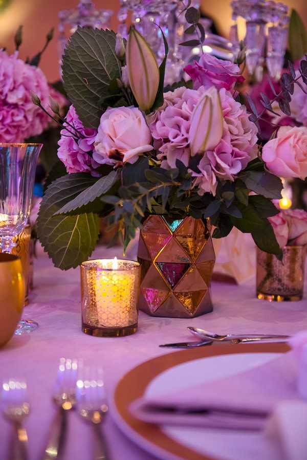 Close-up detail shot of a reception table centerpiece arrangement lit with warm pink ambient lighting. The floral centerpiece features blush pink garden roses, mauve hydrangeas, cream lily buds, and dark green foliage arranged in a geometric faceted vase with copper and jewel-toned stained glass panels in triangular patterns. A mercury glass votive candle holder with a lit candle sits beside the arrangement. The table is set with a white linen, terracotta-rimmed charger plates, silver cutlery, and crystal glassware including what appears to be a chandelier or candelabra in the background. The overall decor palette combines copper, mauve, blush pink, and warm gold tones in a modern yet richly colored styling approach.