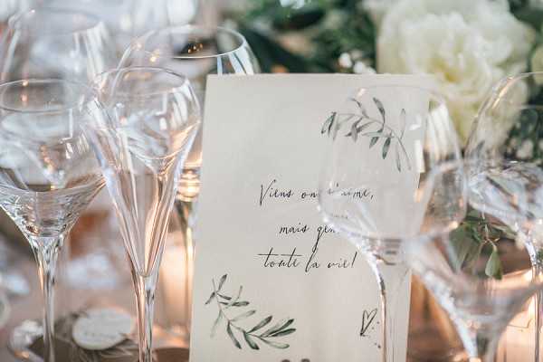 A close-up detail shot of a wedding reception tablescape featuring multiple clear crystal wine glasses arranged around a cream-colored stationery card with handwritten-style script reading 'Viens on s'aime, mais genre toute la vie!' The card is decorated with watercolor botanical illustrations of green leaves and branches. In the background, soft white floral arrangements and warm candlelight create a blurred bokeh effect. The overall decor palette is white, green, and gold with a modern romantic styling theme.