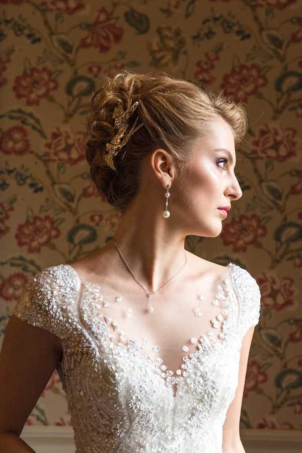 A close-up portrait of a bride photographed in profile against a floral-patterned wallpaper featuring red, green, and cream tones. She wears a heavily beaded and embroidered ivory wedding dress with a sheer illusion neckline, cap sleeves, and intricate floral beadwork across the bodice. Her hair is styled in an upswept chignon adorned with a gold ornamental hair piece, and her accessories include drop pearl earrings and a delicate pearl pendant necklace on a fine chain. The lighting is soft and directional, highlighting her facial features and the texture of the gown's beading.