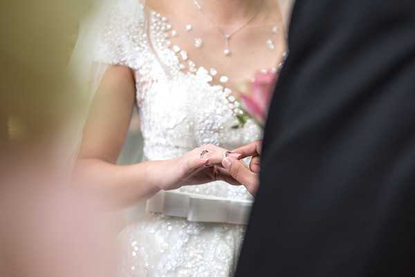 Close-up detail shot of a ring exchange moment during a wedding ceremony, with the groom placing a ring on the bride's finger. The bride is wearing a heavily embellished white lace and beaded cap-sleeve dress with a pearl and crystal necklace, and holds a bouquet with pink flowers partially visible. The groom is dressed in a dark suit. The composition is tightly framed with soft foreground blur framing the hands at center, giving a candid, intimate feel.