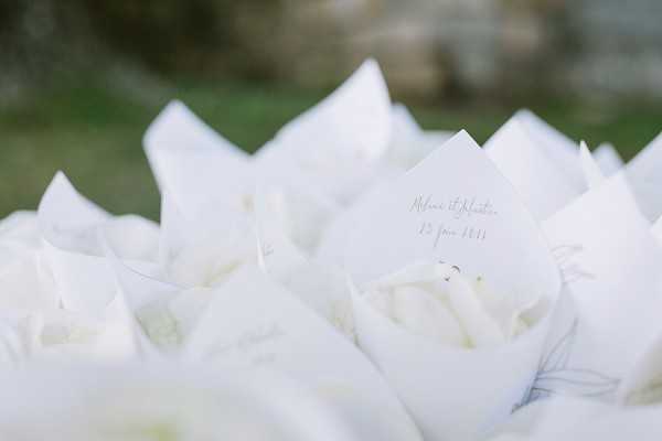 Close-up detail shot of white paper cones filled with white flower petals, arranged together as guest confetti throws. A small white card is visible in the foreground, printed with the couple's names and a date in delicate script lettering. The cones are folded from white paper and styled uniformly, creating a clean, all-white aesthetic. The background is softly blurred with hints of greenery, keeping focus on the stationery detail.