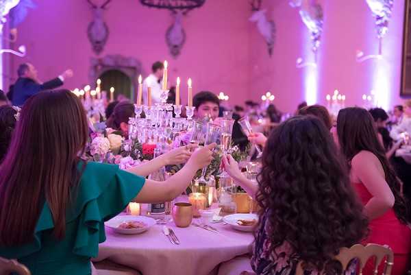 Indoor wedding reception dinner scene captured from a medium shot, showing guests seated at round tables raising champagne glasses in a toast. The ornate ballroom features pink-purple uplighting on decorative wall moldings and archways, creating a warm ambient glow. Table decor includes tall candelabras with white taper candles, crystal glassware stacked in a tower arrangement, white and pink floral centerpieces, gold/copper accent vessels, and votive candles on ivory linen tablecloths. Guests visible include a woman in a teal ruffle-sleeve dress and another in a red dress in the foreground, with a full room of approximately 50 or more attendees visible in the background, suggesting a large-scale formal reception in a chateau or palace-style venue.