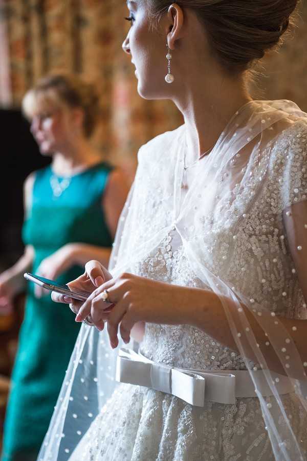 A close-up portrait of a bride indoors, likely during a getting-ready or pre-ceremony moment, holding a smartphone in both hands. She is wearing an ivory sequined lace gown with a white satin bow belt at the waist and a dotted tulle cape or veil draped over her shoulders. Her accessories include drop earrings featuring pearl and crystal details and a pearl ring. Her brown hair is styled in an updo. In the soft background, a woman in a teal sleeveless dress with a necklace is visible, likely a bridesmaid or family member. The setting appears to be an interior room with warm, patterned decor visible in the background.