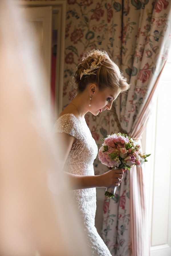 A bridal portrait taken indoors, likely in a getting-ready room, with the bride framed between soft out-of-focus foreground elements suggesting a doorway or curtain. The bride wears a fitted, heavily beaded or sequined white gown with short cap sleeves and looks downward at her bouquet. Her hair is styled in an upswept chignon adorned with a gold ornamental headpiece, and she wears drop pearl earrings. She holds a rounded bouquet of blush pink roses, pale pink freesias, and soft lavender blooms with green foliage. Behind her, floor-length curtains in a classic floral toile pattern in pink, red, and green on a cream background are tied back with a striped blush tie, suggesting a formal interior setting such as a chateau or manor house. The image is a medium portrait shot with a shallow depth of field and warm ambient window light.