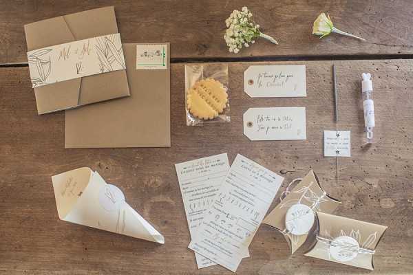 A flat lay arrangement of French wedding stationery and guest favors displayed on a rustic wooden surface. The suite includes a kraft paper pocket-fold invitation with a printed belly band featuring botanical line drawings and handwritten-style calligraphy, a matching kraft envelope, paper confetti cones, luggage-style gift tags with handwritten French text, printed activity or questionnaire cards, a small kraft pillow box tied with twine and a round sticker seal, a packaged shortbread biscuit, a small hand sanitizer or perfume vial, and a white sticker label. A small sprig of baby's breath and a yellow flower bud are placed in the upper right corner as styling accents. The overall palette is warm kraft brown, cream, and white with botanical line-art motifs, suggesting a rustic or natural wedding theme. Wide overhead flat lay composition.