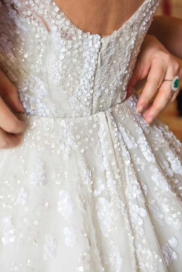 Close-up detail shot of a bride being helped into her wedding dress during the getting-ready portion of the day. The gown is heavily embellished with iridescent white and silver sequins across a tulle base, featuring a fitted bodice with a defined waist seam and a full skirt. A second person's hands are visible fastening the back zipper closure, and they are wearing a statement ring with a large emerald-cut green stone surrounded by a diamond halo. The composition is a tight detail/close-up focusing on the back bodice and waist area of the dress.