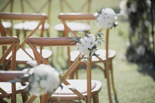 Close-up detail shot of an outdoor wedding ceremony aisle setup featuring natural wood cross-back chairs arranged in rows on a grass lawn. Each chair back is decorated with a small floral arrangement of white flowers — likely ranunculus or lisianthus — with green foliage and olive branch sprigs. The decor palette is white and green, consistent with a classic or garden-style ceremony aesthetic. The background is softly blurred, revealing additional chair rows and what appears to be a floral arch or arrangement in the far right.