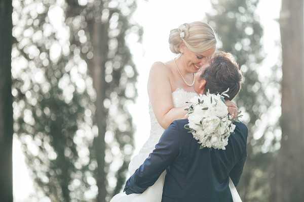 A couple portrait taken outdoors, with the groom lifting the bride as they lean in to kiss. The bride wears a strapless white lace gown with a pearl necklace and a hair accessory, her blonde hair styled in an updo. She holds a bouquet of white peonies and green foliage. The groom wears a navy suit. The background is heavily blurred, showing tall trees with soft backlighting creating a bright, airy atmosphere. The shot is a medium close-up portrait with a shallow depth of field.