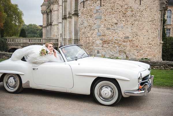 A bride sits in the passenger seat of a vintage white Mercedes-Benz convertible (likely a 190SL) parked on a gravel drive beside a French château with stone masonry and castle turrets visible in the background. The bride wears a voluminous white ball gown with tulle that spills over the car door, and holds a loose, garden-style bouquet with mixed warm-toned blooms and greenery. She is smiling and appears relaxed in a wide portrait-style shot that includes the full length of the classic car. The overall styling is classic and romantic, pairing a heritage vehicle with a traditional bridal look in front of château architecture. Potential venue feature image.