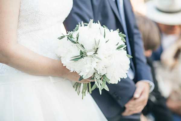 Close-up detail shot of a bride holding a round bouquet composed of white peonies and olive branches with silver-green foliage. The bride is wearing a white lace dress, and a groom in a navy suit is visible in the soft-focus background alongside a guest wearing a white sun hat. The image is taken outdoors, likely during a ceremony, with bright natural light. The bouquet styling is classic and restrained, with a white and green palette.