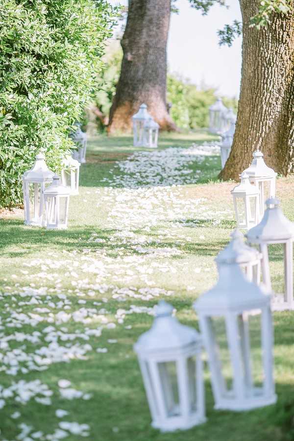 Detail shot of an outdoor wedding ceremony aisle set in a garden or parkland, with no people present. The aisle is lined on both sides with white wooden lanterns of varying heights, arranged in clusters, and scattered white rose petals cover the grass path leading into the distance. The decor palette is entirely white, creating a clean, classic aesthetic. The composition is a perspective shot taken from ground level, with the foreground lanterns in soft focus and the aisle receding toward large trees in the background.