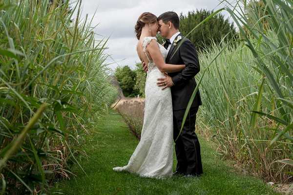 A couple portrait taken outdoors, with the bride and groom standing close together with foreheads nearly touching in an intimate pose. They are positioned along a narrow grass path flanked by tall reeds or ornamental grasses, with a low stone wall visible in the background. The bride wears a fitted ivory lace gown with an open lace back and cap sleeves, while the groom is dressed in a black tuxedo with a white dress shirt and black bow tie. The shot is a medium full-length portrait taken from slightly below eye level, with the tall grasses framing the couple on both sides to create a natural tunnel effect.