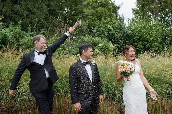 A celebratory outdoor portrait of a bride and groom alongside a male guest who is raising his arm and spraying what appears to be champagne or confetti, all three laughing and reacting joyfully. The setting is an outdoor garden area with tall ornamental grasses and dense green trees in the background. The groom wears a dark tuxedo with a white boutonniere, the male guest wears a black tuxedo with bow tie, and the bride wears a fitted white lace cap-sleeve gown, holding a bouquet of peach and coral roses with greenery. The shot is a medium-wide portrait taken at ground level.