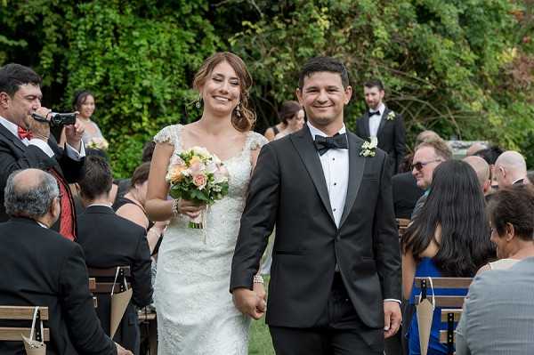 A bride and groom walk back down the aisle together immediately following an outdoor wedding ceremony, holding hands and smiling. The bride wears a fitted, cap-sleeve lace gown in ivory and carries a bouquet of peach and blush roses with greenery, while the groom wears a dark charcoal suit with a black bow tie and a white boutonniere. Seated guests line both sides of the aisle on wooden chairs, dressed in formal attire, with a musician holding a microphone visible on the left side. The setting is an outdoor garden ceremony with lush green trees in the background, captured in a mid-range portrait shot.