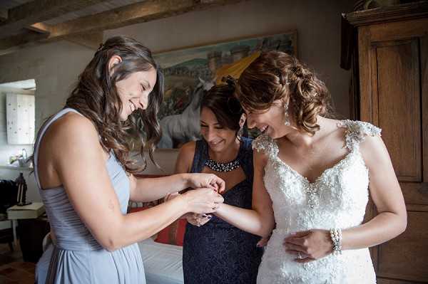 A getting-ready moment captured indoors, showing the bride and two women — one in a pale blue-grey sleeveless gown and one in a dark navy dress with a statement necklace — examining or fastening jewelry on the bride's wrist, all three smiling and laughing. The bride wears an ivory lace cap-sleeve gown with beaded detailing and a pearl bracelet, with her hair styled in an updo. The room features rustic exposed wooden ceiling beams, a large wooden armoire, and a colorful painting on the wall, suggesting a French farmhouse or chateau interior. The shot is a medium portrait taken at close range, with warm natural light coming from the left side of the frame.