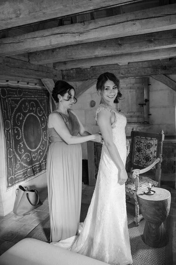 A black-and-white getting-ready shot of a bride and one bridesmaid inside a rustic interior space with exposed wooden ceiling beams and stone or plaster walls. The bridesmaid, wearing a floor-length draped gown, is fastening the back of the bride's fitted lace wedding dress with a cap-sleeve detail, while the bride turns her head toward the camera with a smile. The room features an ornate embroidered textile hanging on the wall, a carved wooden chair with a patterned seat, a small wooden stool holding a candle, and a leather tote bag on the floor, giving the space a bohemian, rustic character. The image is a medium full-length portrait with soft natural light coming from the left side, creating moderate contrast between the bright dress and the darker background tones.