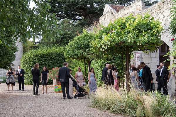 Wedding guests mingle during what appears to be a cocktail hour in an outdoor courtyard or garden area adjacent to a historic stone building with arched architectural details and ivy-covered walls. Approximately 20 guests are visible in small conversational groups spread across a gravel pathway, with one guest pushing a black stroller. Guests are dressed in formal attire, with women wearing dresses in shades of grey, silver, and black, and men in dark suits. The setting is landscaped with trimmed hedgerows, ornamental grasses, and trained trees forming a natural canopy over part of the gathering space. Wide shot capturing the full scene and architectural context. Potential venue feature image.