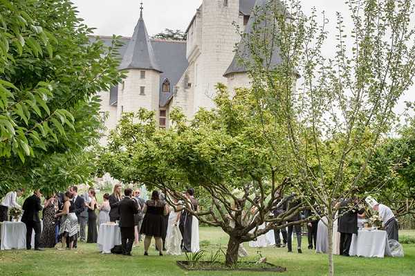 A cocktail hour takes place on the lawn grounds of a French chateau, with approximately 30 guests mingling in small groups across a wide garden area. The chateau visible in the background features pale stone construction with dark slate-roofed turrets and Gothic-style windows. Guests are dressed in formal attire including dark suits and black dresses, with the bride visible in a white gown near the center-right of the frame. White-clothed cocktail tables are positioned throughout the lawn, and at least one table appears to be set with food or a display. The wide shot is taken from a low angle framed by tree branches in the foreground, giving depth to the scene. Potential venue feature image.