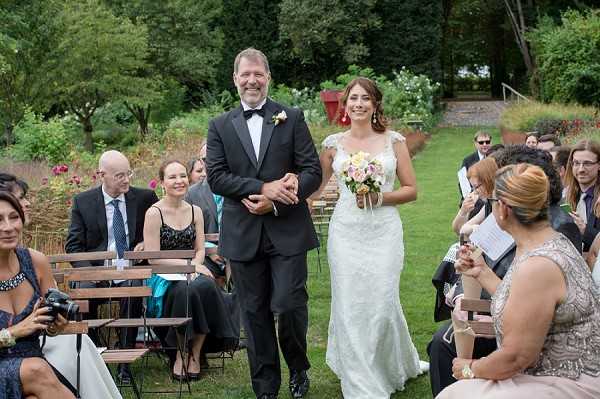 An outdoor garden wedding ceremony showing a bride being walked down the aisle by a male companion, presumed to be her father. The bride wears a fitted white lace gown with cap sleeves and carries a bouquet of pale pink and white blooms with greenery. Her escort is dressed in a black tuxedo with a bow tie and a small boutonniere. Seated guests line both sides of the wooden folding chair aisle, dressed in formal attire including a woman in a black dress and another in a teal outfit. An officiant in a silver beaded dress stands to the right holding papers. The setting is a lush garden with mature trees and shrubs in the background. Wide shot taken from near the front of the aisle looking back toward the couple.