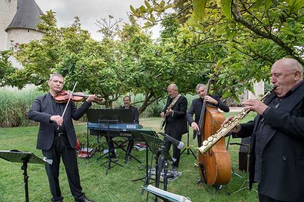 A five-piece live band performs outdoors in a garden setting, likely during a cocktail hour or reception. The musicians, all dressed in black suits, are playing violin, keyboard (Korg), guitar, double bass, and saxophone. The group is set up on a lawn with a chateau tower visible in the background. The shot is a wide, candid performance photo taken at ground level.