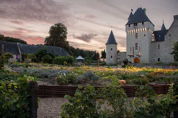 Wide-angle exterior shot of a French château at dusk, featuring a multi-turreted stone castle with grey slate conical roofs and lit windows visible on the right, alongside a lower stone outbuilding with a grey roof on the left. In the foreground, raised wooden garden beds contain lush plantings including what appears to be a vegetable and herb potager garden, with grapevines along a wooden fence bordering a gravel path. A white event marquee is visible in the middle ground between the outbuilding and the château towers, suggesting a wedding reception setup. No people are visible in the frame. Potential venue feature image.