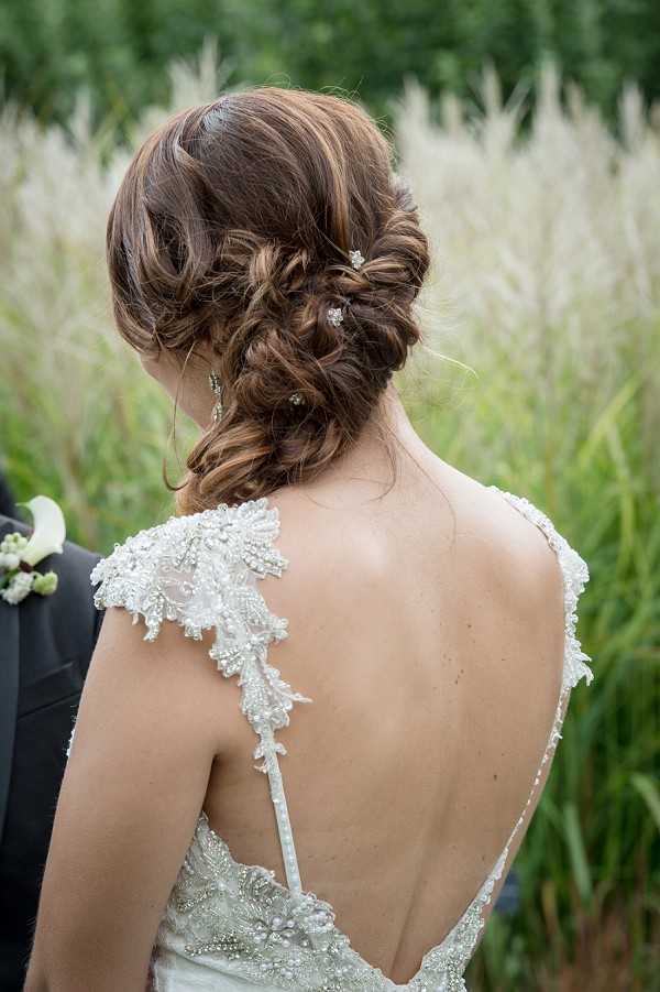 Close-up detail portrait of a bride shot from behind, outdoors with tall ornamental grasses visible in the background. The image focuses on the bride's low twisted updo hairstyle, which is decorated with small crystal hair pins, paired with drop crystal earrings. She is wearing a low-back ivory gown with heavily beaded and embroidered lace cap sleeves and a deep V-back neckline with a thin spaghetti strap detail. A portion of a groom in a dark suit with a white floral boutonniere is partially visible on the left edge of the frame.