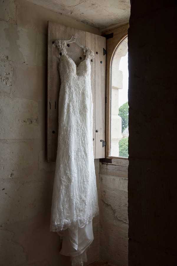 A detail shot of a wedding dress hanging from a wooden shutter beside an arched stone window inside what appears to be a historic chateau or castle. The dress is a fitted, floor-length ivory gown with all-over lace and beading, a sweetheart neckline, and thin lace-edged straps. The interior walls are rough-cut stone, and natural light filters in through the arched window, illuminating the dress against the rustic backdrop.