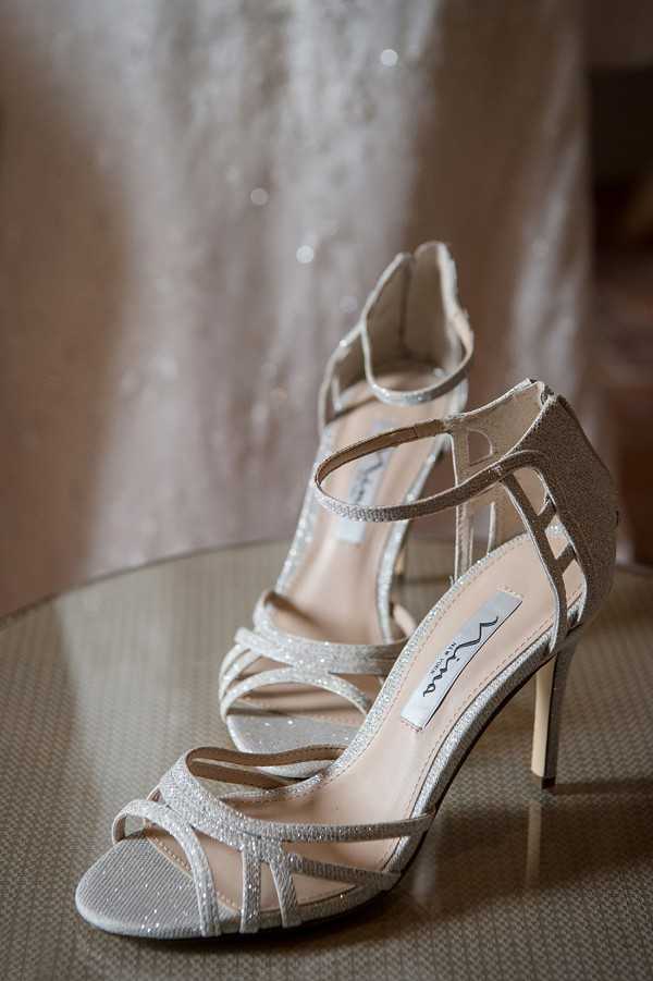 A close-up detail shot of a pair of silver glitter strappy stiletto heels placed on a reflective glass surface. The shoes feature a cage-style multi-strap design with an open toe and ankle strap, and a branded insole label is visible inside. In the soft-focus background, a beaded or lace wedding dress is partially visible, suggesting this is a bridal getting-ready scene. The overall styling is classic and polished, with the metallic silver shoes as the clear focal point.