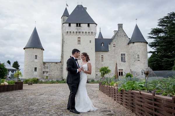 A couple poses together on a cobblestone courtyard in front of a large French château featuring multiple conical-roofed towers and pale stone architecture. The groom wears a classic black tuxedo with a bow tie, while the bride wears a fitted, sleeveless white gown with a slight train. They stand close together, facing each other in a relaxed portrait pose. Raised wooden planter beds with vegetable garden plantings frame the foreground of the courtyard. This is a medium wide shot capturing both the couple and the full façade of the château behind them. Potential venue feature image.