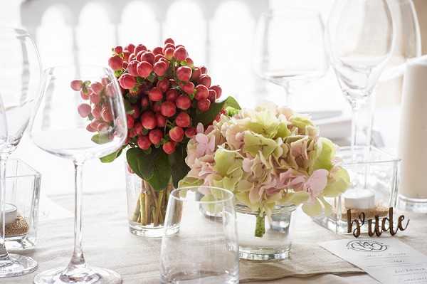 Close-up detail shot of a wedding reception table setting featuring two small floral arrangements in clear glass vases: one composed of red hypericum berries with green leaves, and one of blush pink and sage green hydrangeas. A laser-cut wooden 'bride' place card marker sits beside the arrangements, accompanied by clear glassware including wine glasses and a tumbler, a small candle, and a printed menu card on a neutral linen tablecloth. The styling palette is soft and romantic, combining red berry tones with muted blush and green florals against a bright, airy indoor setting. The composition is a shallow depth-of-field detail shot with the background softly blurred.